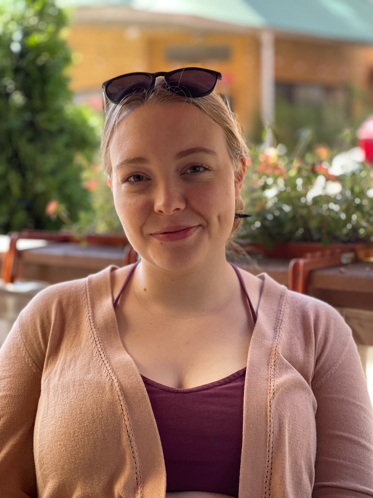 A fair skinned, blonde young woman with her hair pulled back in a bun sits outside, smiling.